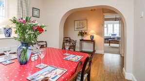 A dining area with a red tablecloth, wooden chairs, and a vase with flowers. There is a framed picture on the wall and a lamp on a side table. - The Pink House (Rhosneigr)