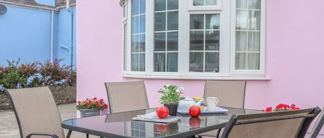 A round table with four chairs, a potted plant, and a teapot on a patio against a pink wall with a bay window.