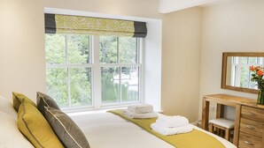 A bedroom with a large bed, a window with a view of trees and water, a wooden dresser, and a mirror. - Gilson (Ambleside)