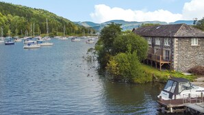 A lakeside property with a stone building, docked boats, and a forested hillside. - Gilson (Ambleside)