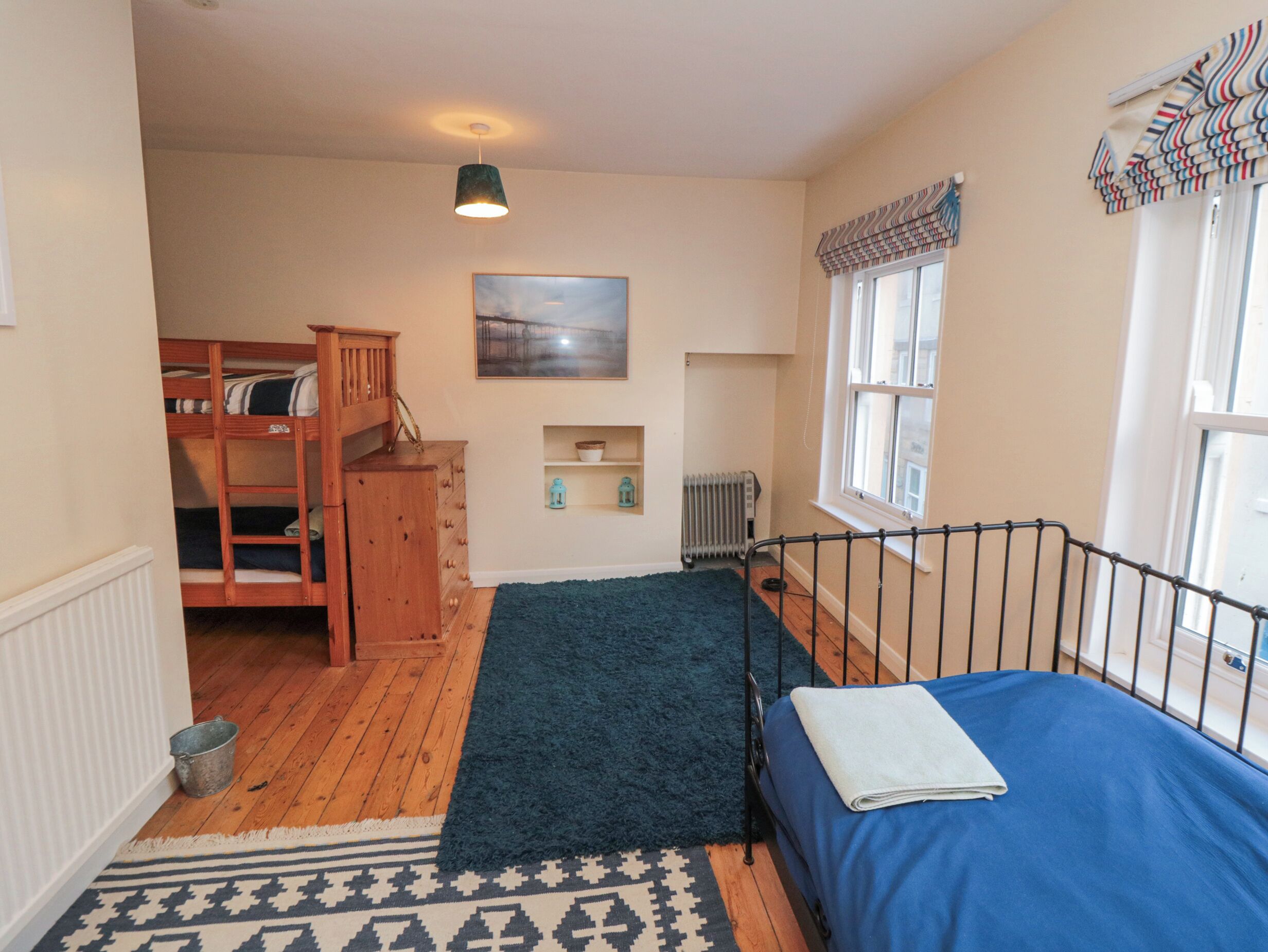 Un dormitorio con literas, una cama individual, un radiador, una ventana con cortinas, un cuadro en la pared y un estante pequeño con objetos decorativos.