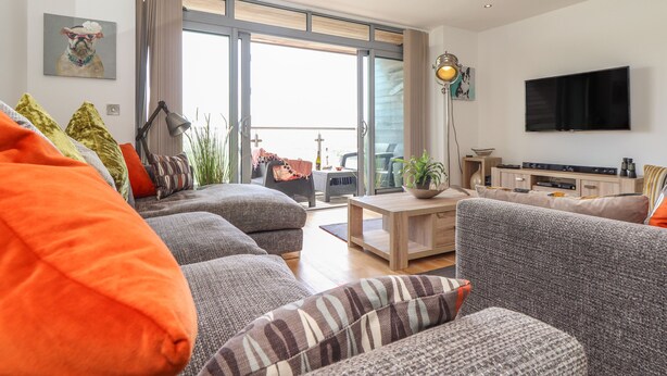 A modern living room with a large TV, comfortable seating, and a balcony with outdoor furniture. - 8 Cribbar (Newquay)