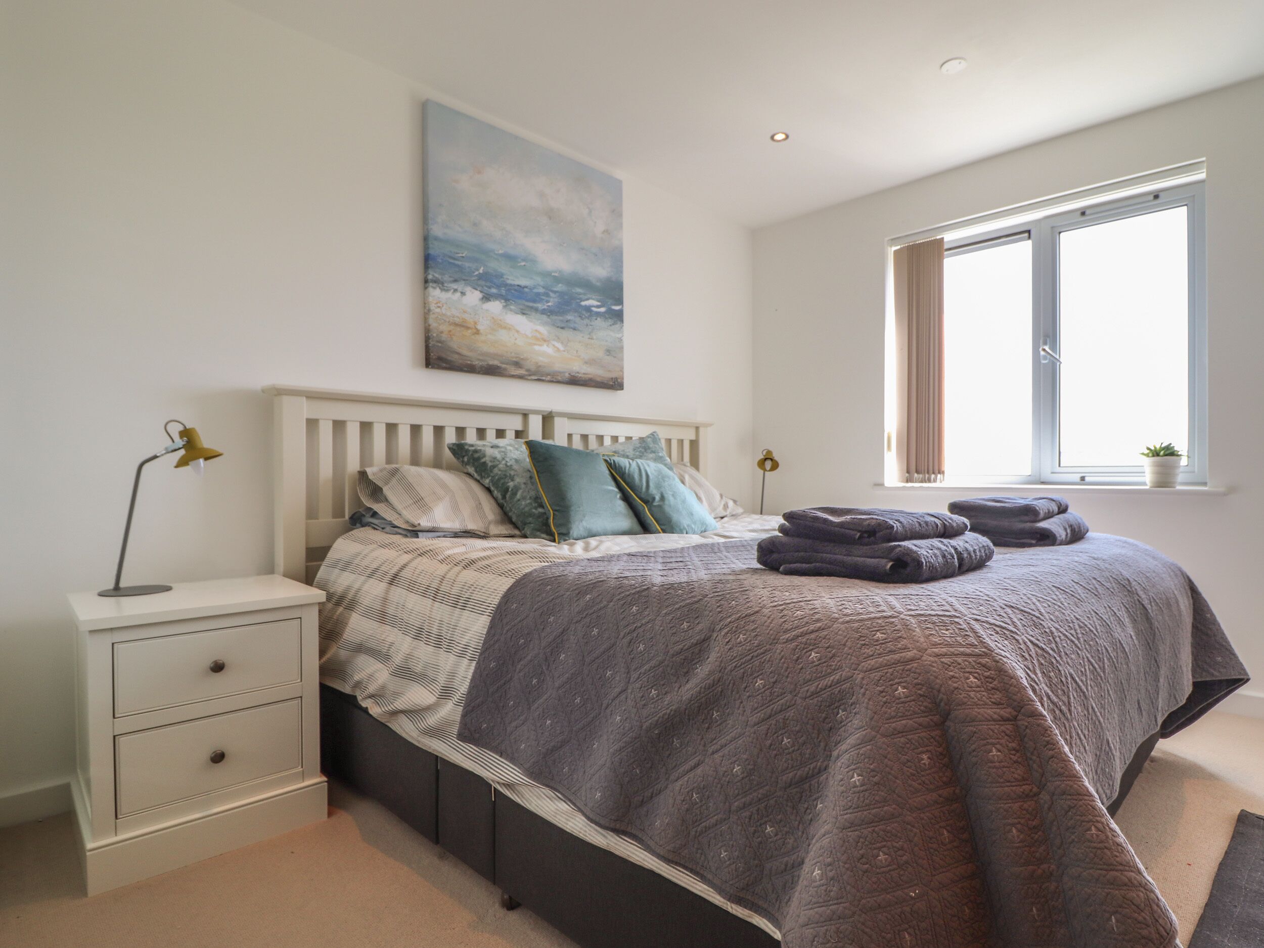 A bedroom with a bed, bedside tables, a painting, a window, and a potted plant.