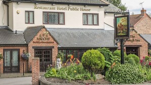 Un bâtiment portant l’inscription « Restaurant Hôtel Bar », de conception traditionnelle, avec une façade en briques, un toit en tuiles et un petit jardin agrémenté de diverses plantes et fleurs.