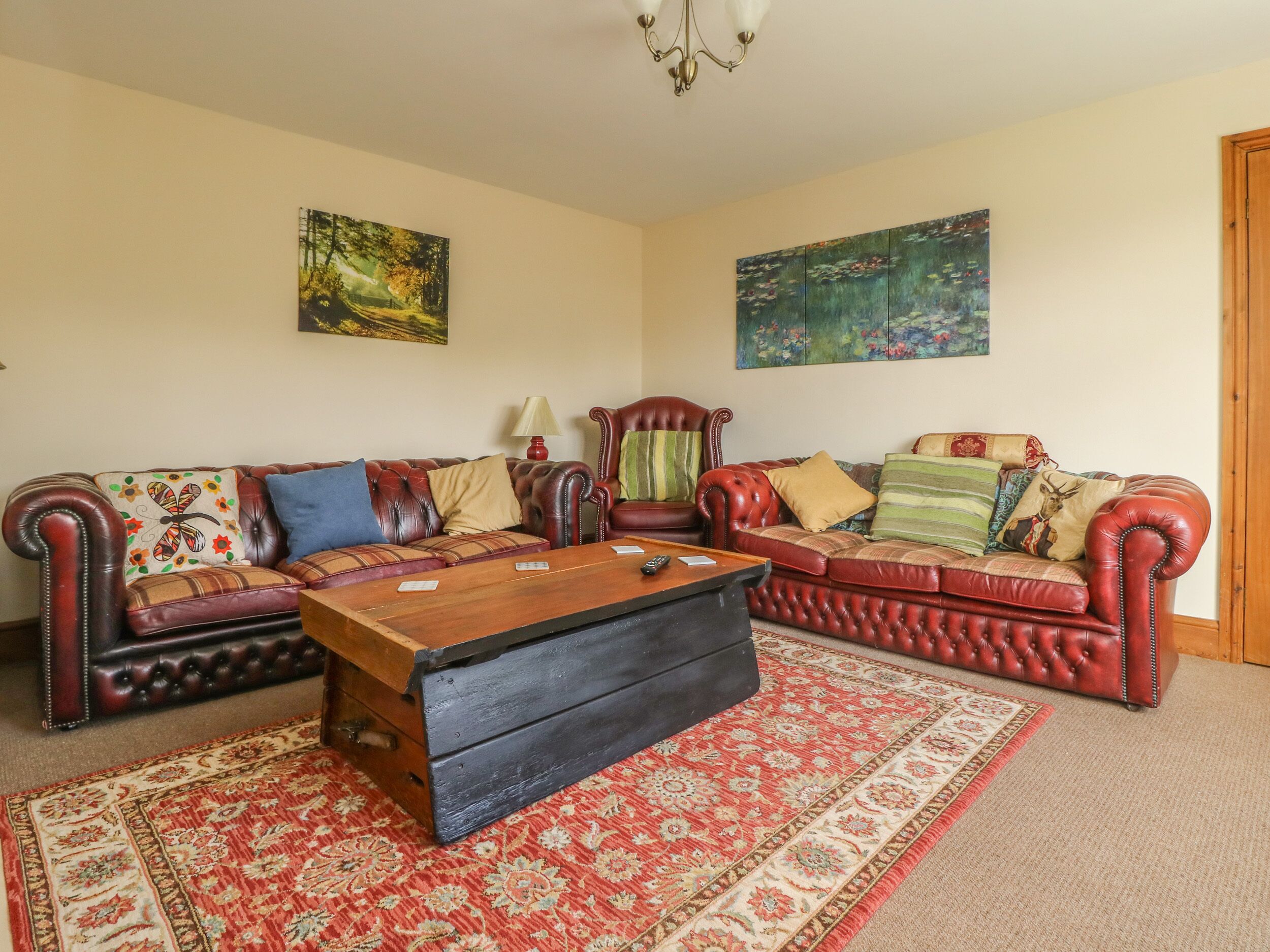 Un salon avec un canapé en cuir rouge, une table basse en bois et un tapis à motifs. Deux tableaux ornent les murs, et un lustre est suspendu au plafond.
