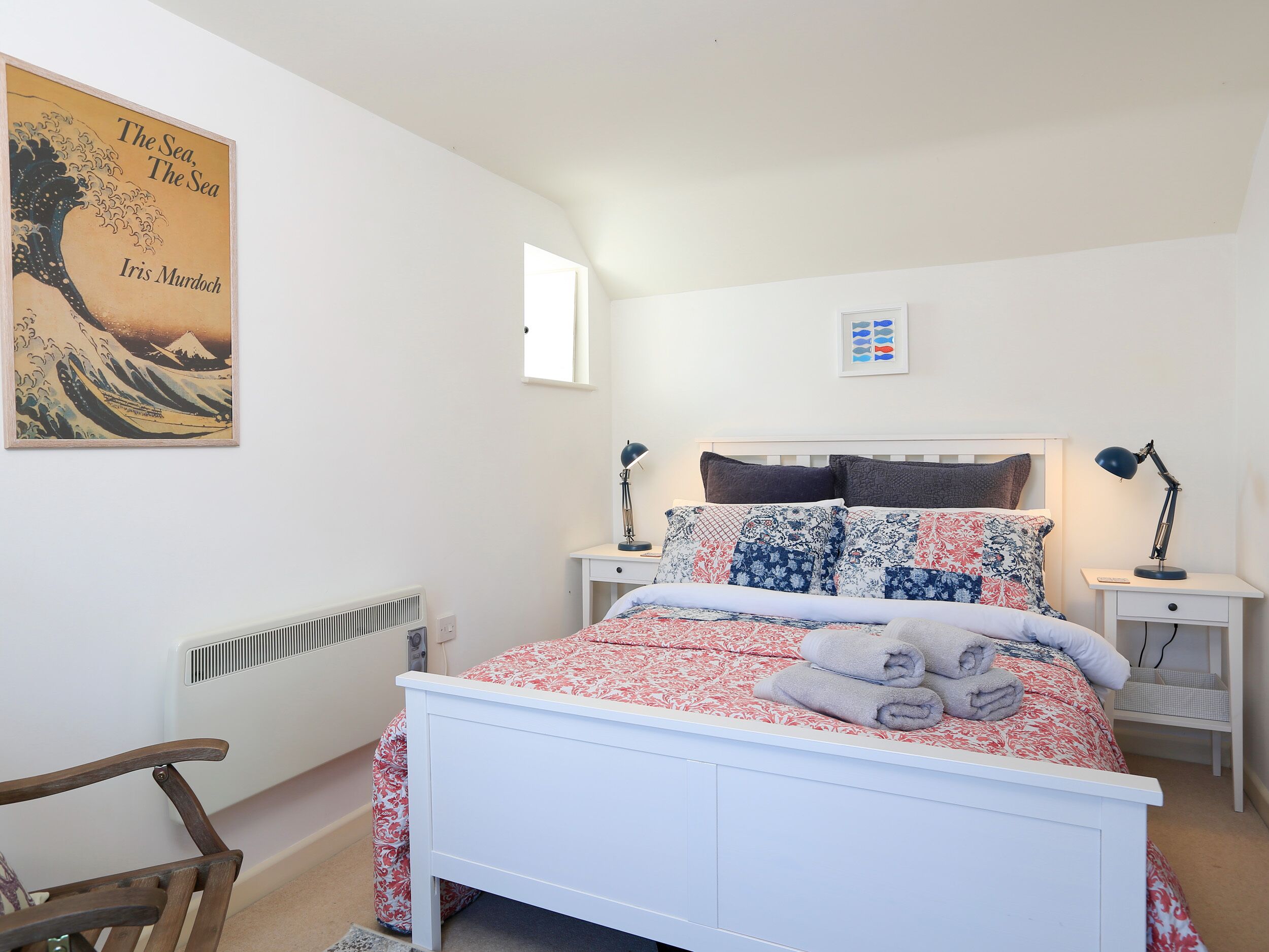A bedroom with a white bed, patterned bedding, a chair, a bedside table with a lamp, and a wall-mounted picture.