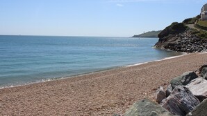 A pebbled beach with a rocky shoreline, a clear sky, and a distant lighthouse. - The Reach (Kingsbridge)