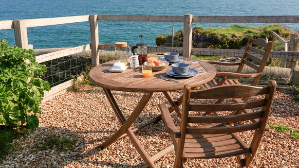 A wooden table with a view of the sea, a chair, and a breakfast setup including a jug, cups, and a plate of food. - The Reach (Kingsbridge)