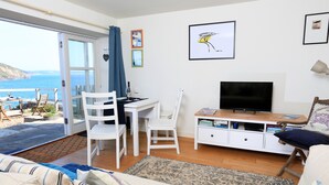 A living room with a sofa, a TV, a dining table with chairs, a bookshelf, and a view of the sea. - The Reach (Kingsbridge)