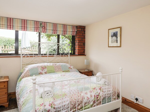 A bedroom with a white metal bed frame, floral bedding, a window with a striped valance, a wooden nightstand with a lamp, and a framed picture on the wall.