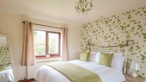 A bedroom with a bed, a window with curtains, a mirror, a nightstand with a lamp, and floral wallpaper. - Berrys Place Farm Cottage (Gloucester)