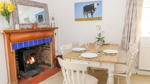 Une salle à manger avec une cheminée, des chaises blanches, une table en bois dressée pour quatre personnes et un tableau représentant une vache accroché au mur.