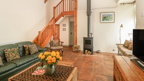 Un salón amplio con techo alto, vigas de madera, una escalera, una chimenea, un sofá verde, una mesa de centro de madera con un jarrón de flores y un televisor.