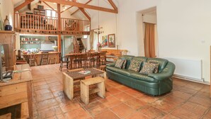 Un salón amplio con techo alto y vigas de madera a la vista, un sofá de cuero verde, muebles de madera y una cocina al fondo.