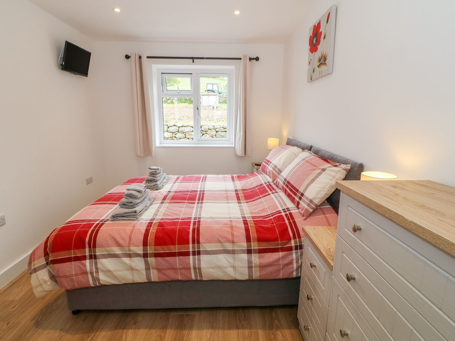 A bedroom with a bed, a nightstand, a dresser, a window with curtains, and a wall-mounted TV.