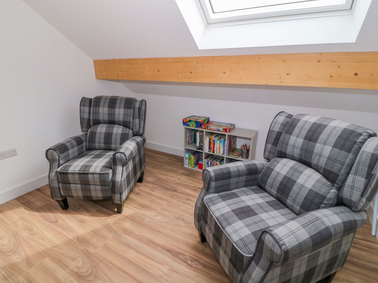 Une pièce avec deux fauteuils à motifs écossais, un plafond avec une poutre en bois, une lucarne et une bibliothèque remplie de livres et de jouets.
