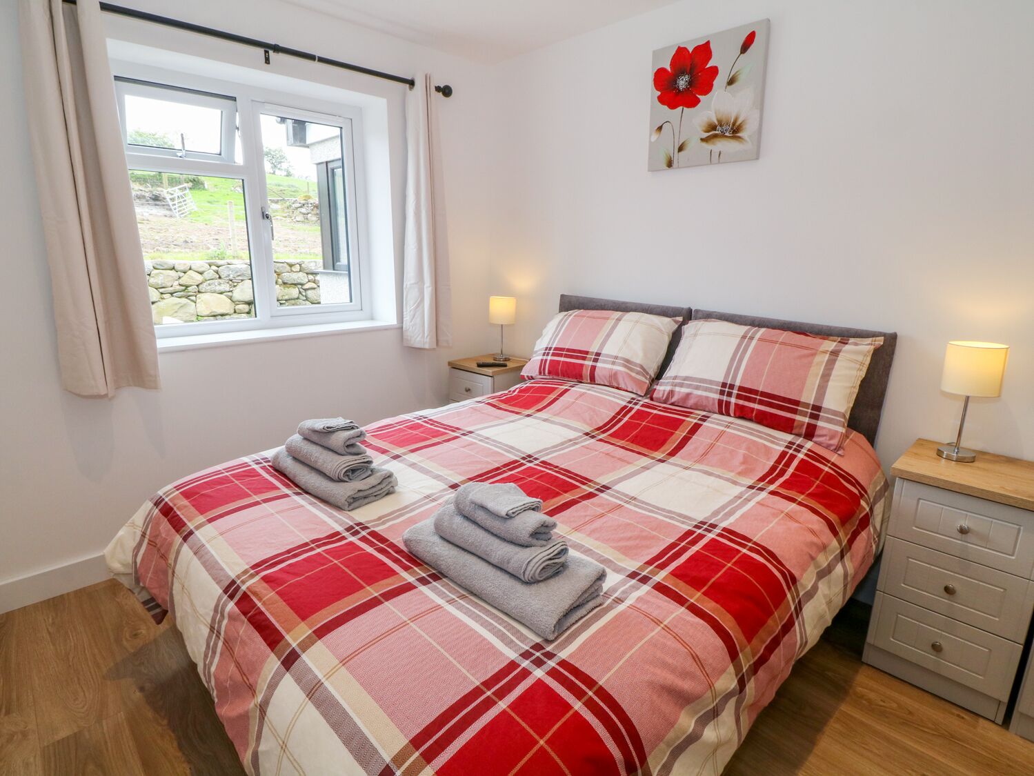 A neatly made bed with a plaid bedspread, two bedside lamps, a small bedside table, and a window with curtains.