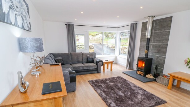 A modern living room with a sofa, a wooden desk, a fireplace, and a view through large windows. - Llwyd Coed Canol (Caernarfon)