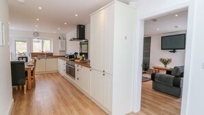 A modern kitchen with white cabinets, a dining area with a wooden table and chairs, and a living room with a sofa and a flat-screen TV. - Llwyd Coed Canol (Caernarfon)