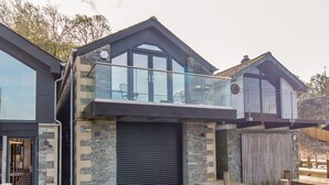 A modern house with a stone facade, a glass balcony, and a wooden deck over water. - The Boat House at Louper Weir (Windermere)