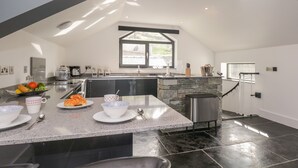 A modern kitchen with a stone island, a dining area with a table and chairs, and a view of the outdoors through a window. - The Boat House at Louper Weir (Windermere)