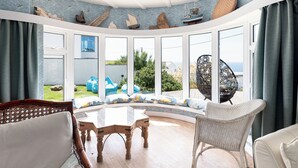 A conservatory with a curved glass wall, wooden furniture, and a view of the sea. - Blue Bay Beach House (Newquay)