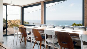 A dining area with a long table, several chairs, and large windows offering a view of the sea. - Blue Bay Beach House (Newquay)