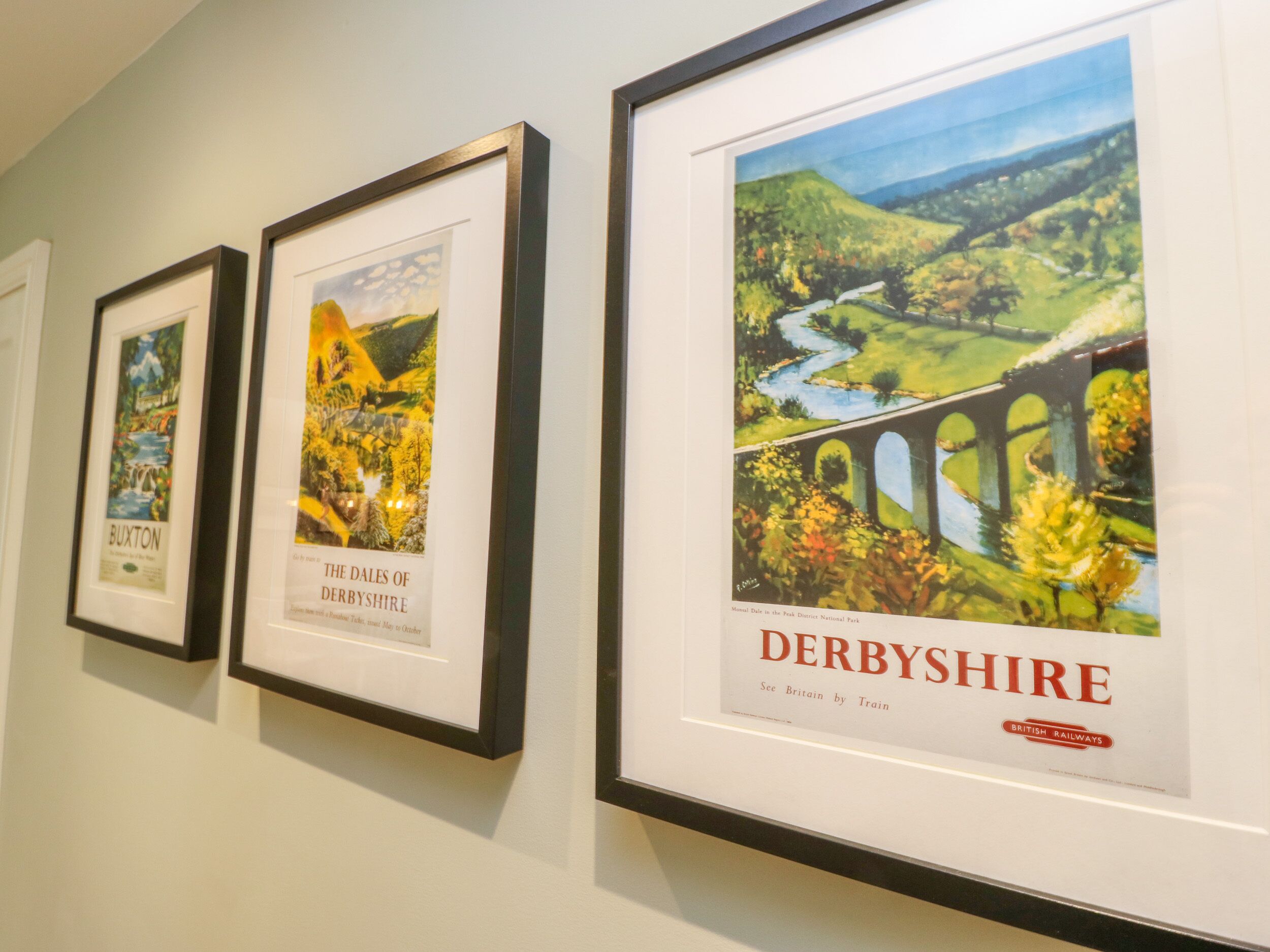 Three framed posters on a wall, each depicting different landscapes and labeled with the names of places: Buxton, The Dales of Derbyshire, and Derbyshire.
