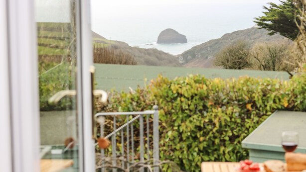 A view from a window overlooking a garden with a table and chairs, a glass door, and a view of the sea and hills. - Glenhaven (Tintagel)