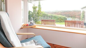 A room with a view of a hillside, a wooden chair, a small table with a tray of croissants, and a window with a curtain. - Glenhaven (Tintagel)