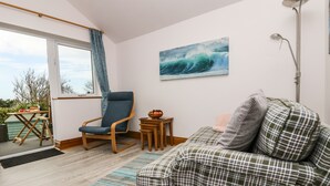 A cozy living room with a sofa, a chair, a small table with a bowl of fruit, and a painting of waves on the wall. - Glenhaven (Tintagel)