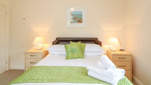 A neatly made bed with a green bedspread, two bedside lamps, a framed picture on the wall, and a wooden nightstand with a drawer. - Forget-me-not (St. Columb Major)