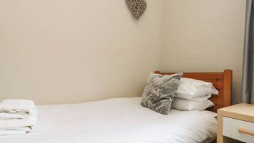 A single bed with white linens, a wooden headboard, a gray patterned pillow, and a small bedside table with a drawer.