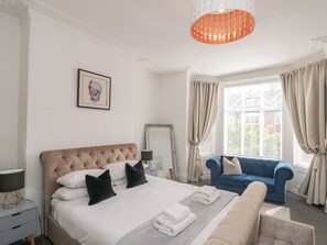 A hotel room with a bed, a blue armchair, a bedside table with a lamp, and a window with curtains. - Royal Victoria House (Scarborough)