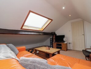 Une pièce avec un plafond en pente, un puits de lumière, un téléviseur, un ventilateur, une table en bois et des sièges de couleur orange.