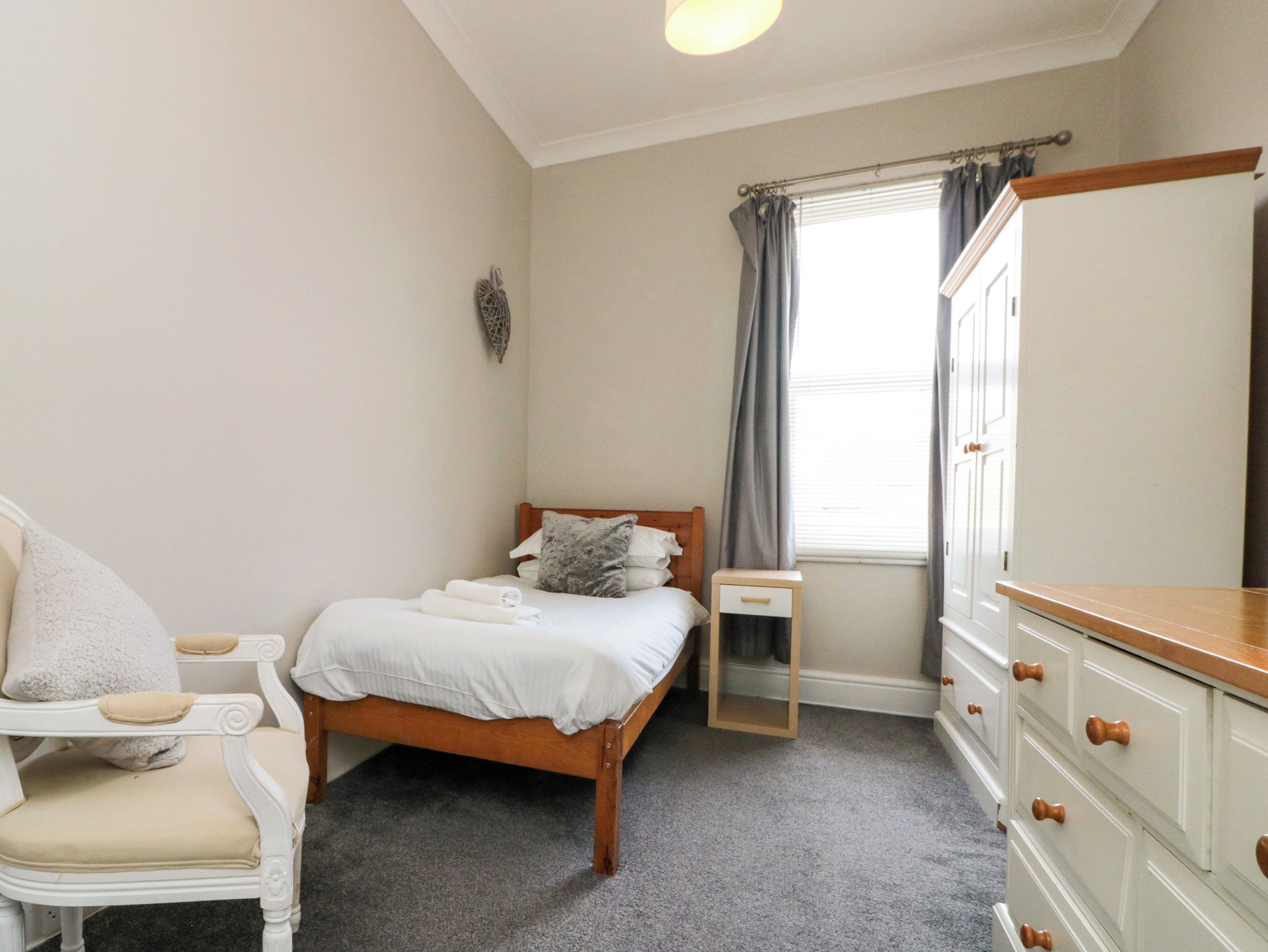 Un dormitorio con cama, una silla, un aparador y una ventana con cortinas.