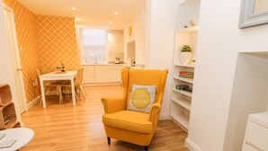 A compact living space with a yellow armchair, a dining table with chairs, a kitchen area, and a bookshelf. - West Cliff Apartment (Whitby)