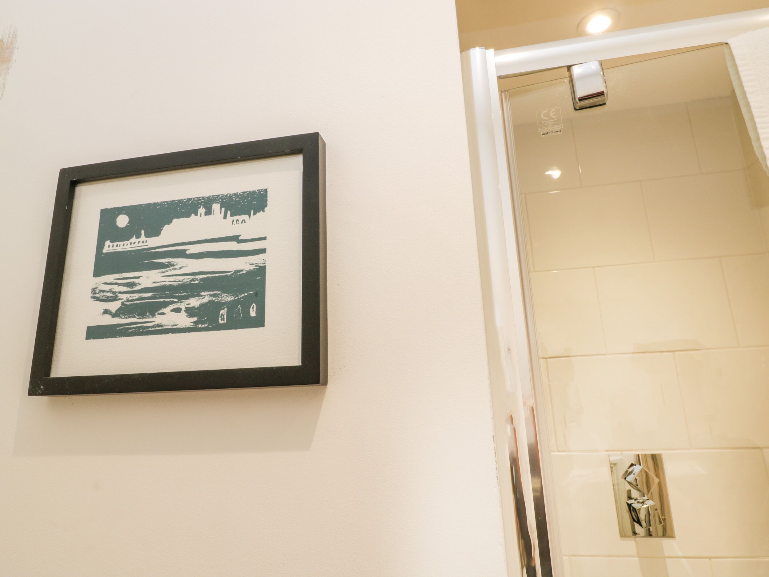 A framed picture of a coastal scene on a wall, a shower area with a glass door, and a tiled wall.