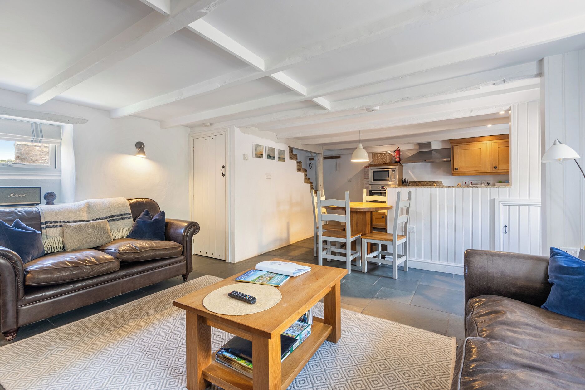 Een woonkamer met een bruine leren bank, een houten salontafel met boeken, en een kamerbreed tapijt. Er is een eethoek met een houten tafel en stoelen, en een keuken met witte kasten en een hangende lamp.