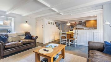 Een woonkamer met een bruine leren bank, een houten salontafel met boeken, en een kamerbreed tapijt. Er is een eethoek met een houten tafel en stoelen, en een keuken met witte kasten en een hangende lamp.