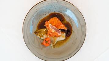 Ein Teller mit einer Portion Essen, möglicherweise Fisch, bedeckt mit Semmelbröseln und einem Stück Fleisch, serviert mit dunkler Sauce und Gemüse.