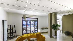 A modern interior with a yellow sofa, a black shelving unit, and a large window with a view of the outdoors. - Hotel Lido Beach (Oristano)
