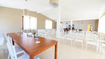 Une pièce spacieuse avec une longue table en bois et des chaises blanches, de grandes fenêtres et un plafond haut.