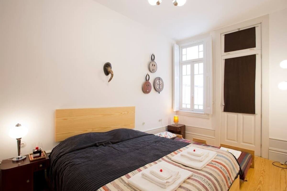 Une chambre à coucher avec une tête de lit en bois, un lit recouvert d’une couvre-lit à rayures, une table de chevet avec une lampe et une fenêtre avec des stores.