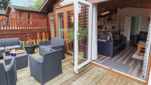 Eine Holzterrasse mit Korbmöbeln, eine Glastür zu einem Wohnbereich mit einer Couch und einem Esstisch.