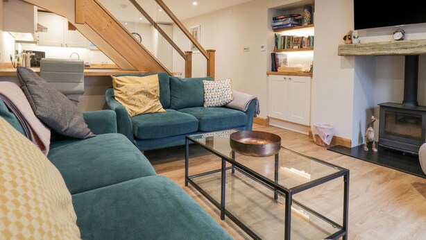 A modern living room with a teal sofa, a glass coffee table, a wooden staircase, a kitchen area, and a fireplace.