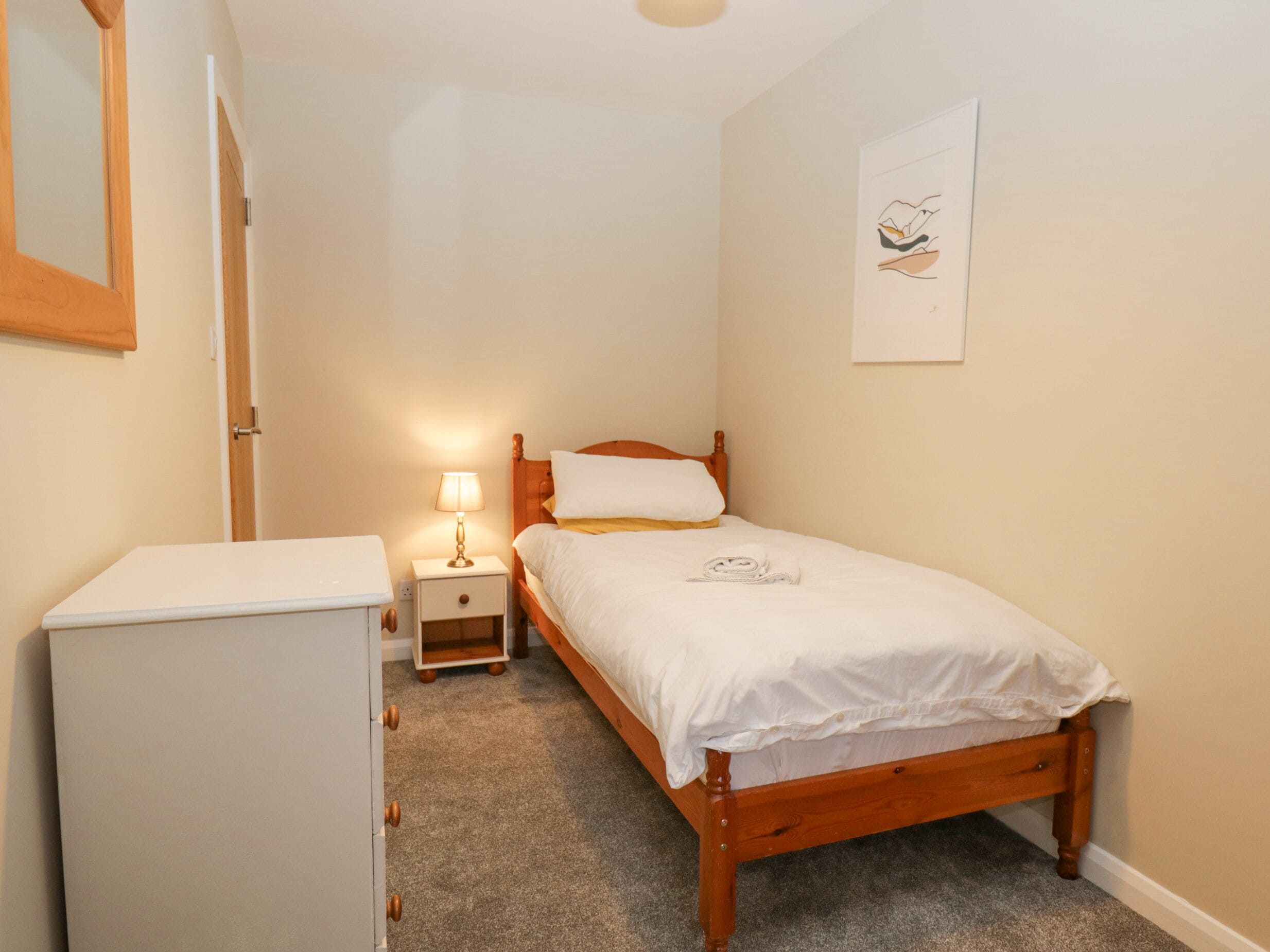 A bedroom with a wooden bed, a nightstand with a lamp, a white dresser, and a framed picture on the wall.