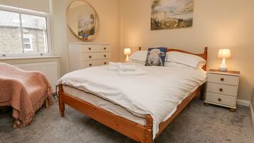 Un dormitorio con cama de madera, ropa de cama blanca, una mesita de noche con lámpara, cómoda, espejo y un cuadro en la pared.