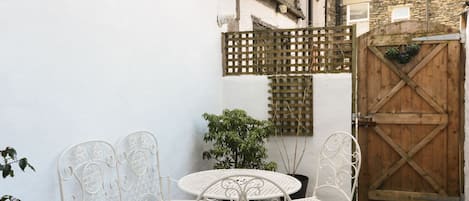 Een binnenplaats met witte metalen stoelen en een tafel, een houten poort en een plant in een pot.