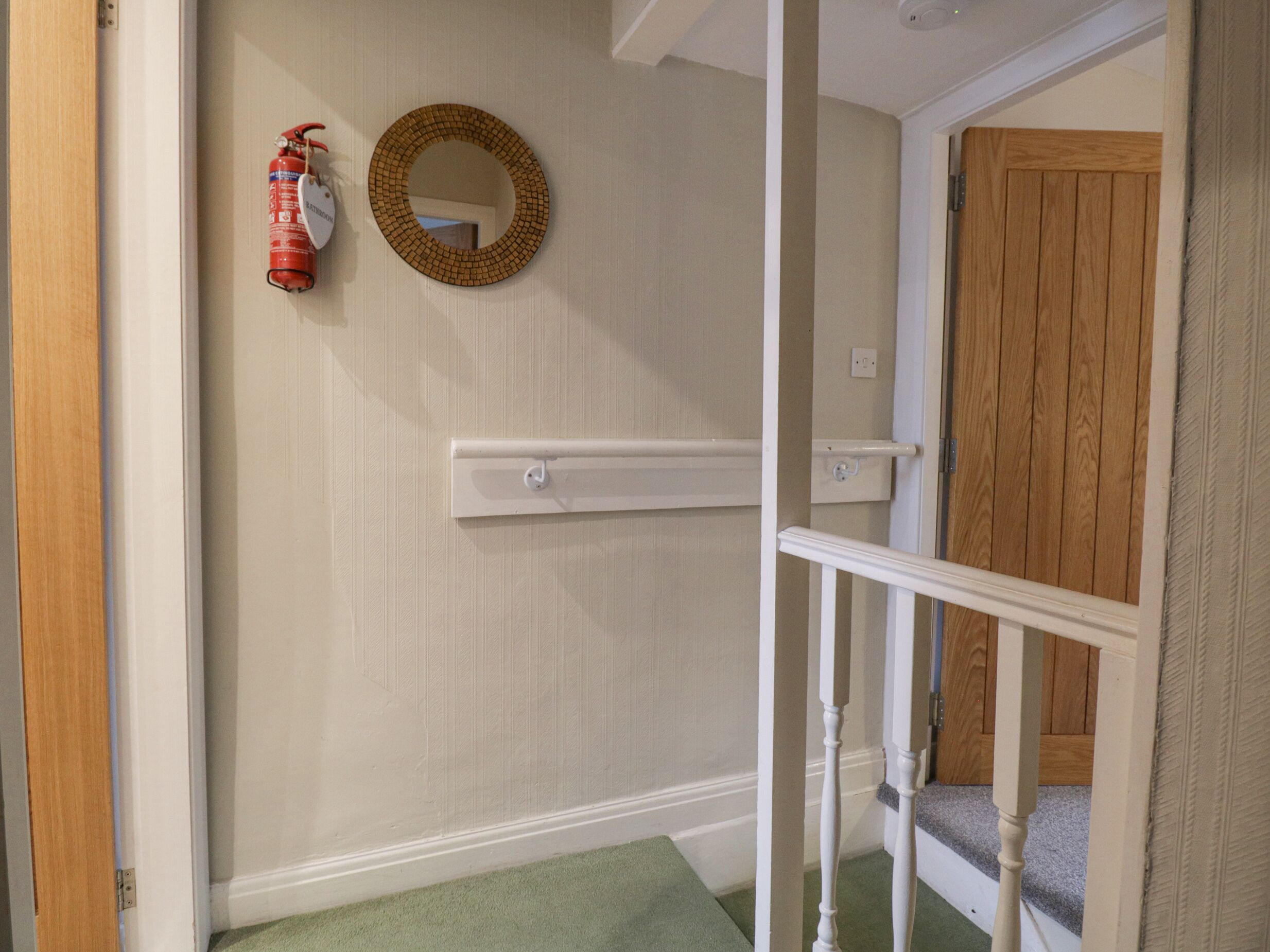 Un pasillo con una puerta de madera, una pared blanca, un espejo y un extintor.